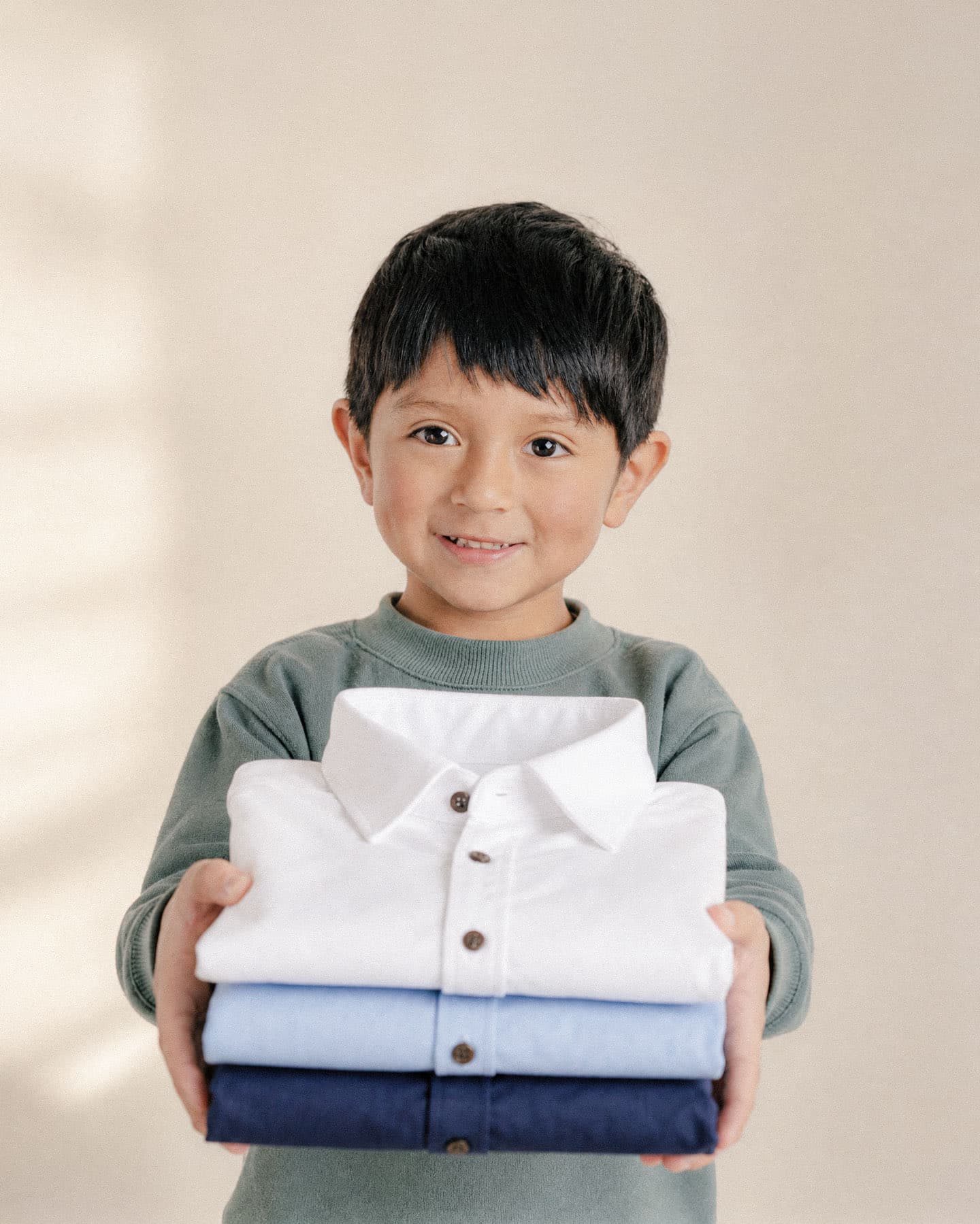 Niño sonriente sosteniendo tres camisas dobladas, blanca, celeste y azul marino, sobre un fondo cálido beige